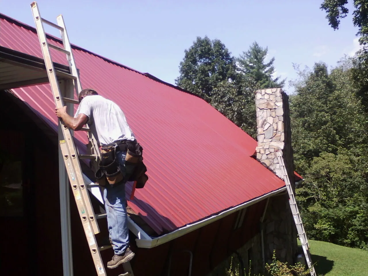 Roofer on a ladder beside a red metal roof and stone chimney — Soffit & Fascia Repair by Roofing Brothers in Stout